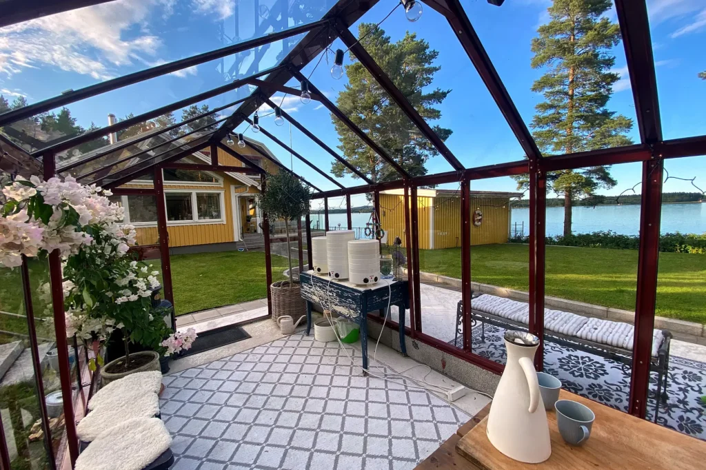 Stormsäkert växthus Primus 249 med mur 20 cm i röd lackering i en svensk villaträdgård med utsikt över vattnet